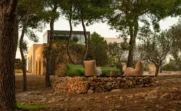 Cozy outdoor seating area surrounded by trees and stone walls, featuring green cushions and a natural setting. Modern house in the background adds a contemporary touch to the serene landscape.
