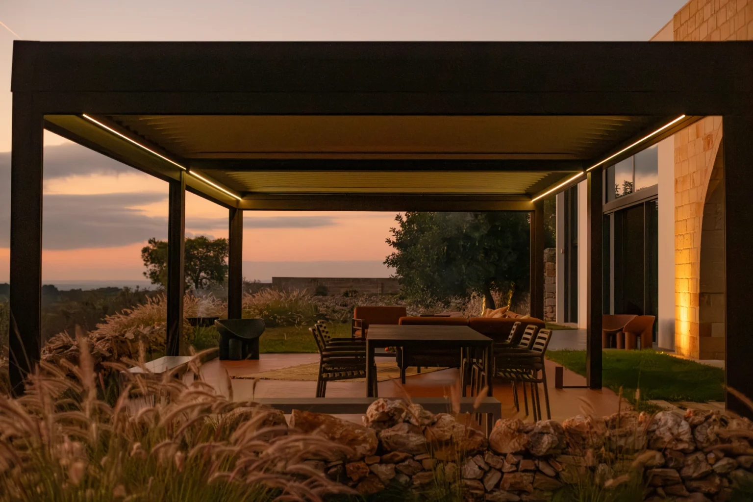 Modern outdoor patio with a sleek pergola, featuring a dining table and chairs, surrounded by lush greenery and a sunset view. Ideal for outdoor entertaining and relaxation. | Only on SopranoVillas