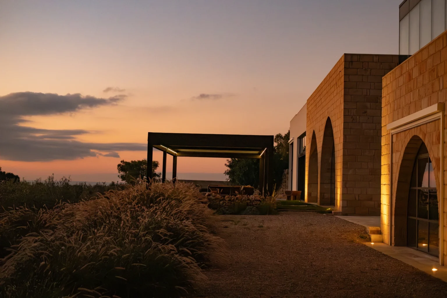 Modern stone building with arched windows and outdoor seating area, surrounded by lush greenery at sunset. | Only on SopranoVillas