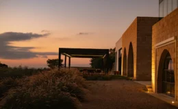 Modern stone building with arched windows and outdoor seating area, surrounded by lush greenery at sunset.