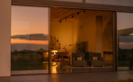 Modern living room with large glass windows reflecting a sunset, showcasing comfortable seating and warm lighting, creating a cozy ambiance.