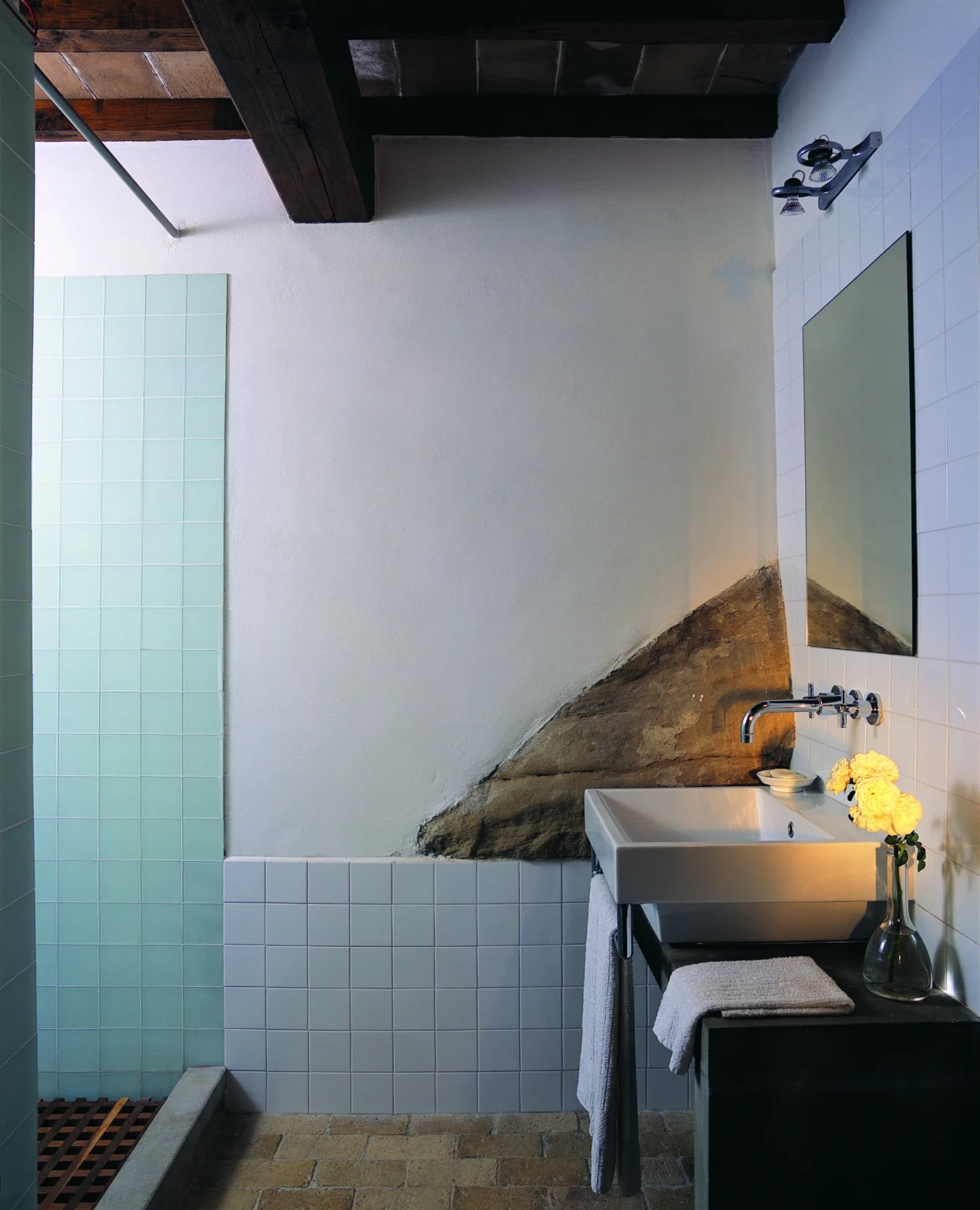 Modern bathroom design featuring a minimalist sink, a large mirror, and a unique textured wall with natural elements. The shower area is separated by glass tiles, creating a sleek and contemporary look. Yellow flowers add a touch of color to the space. | Only on SopranoVillas