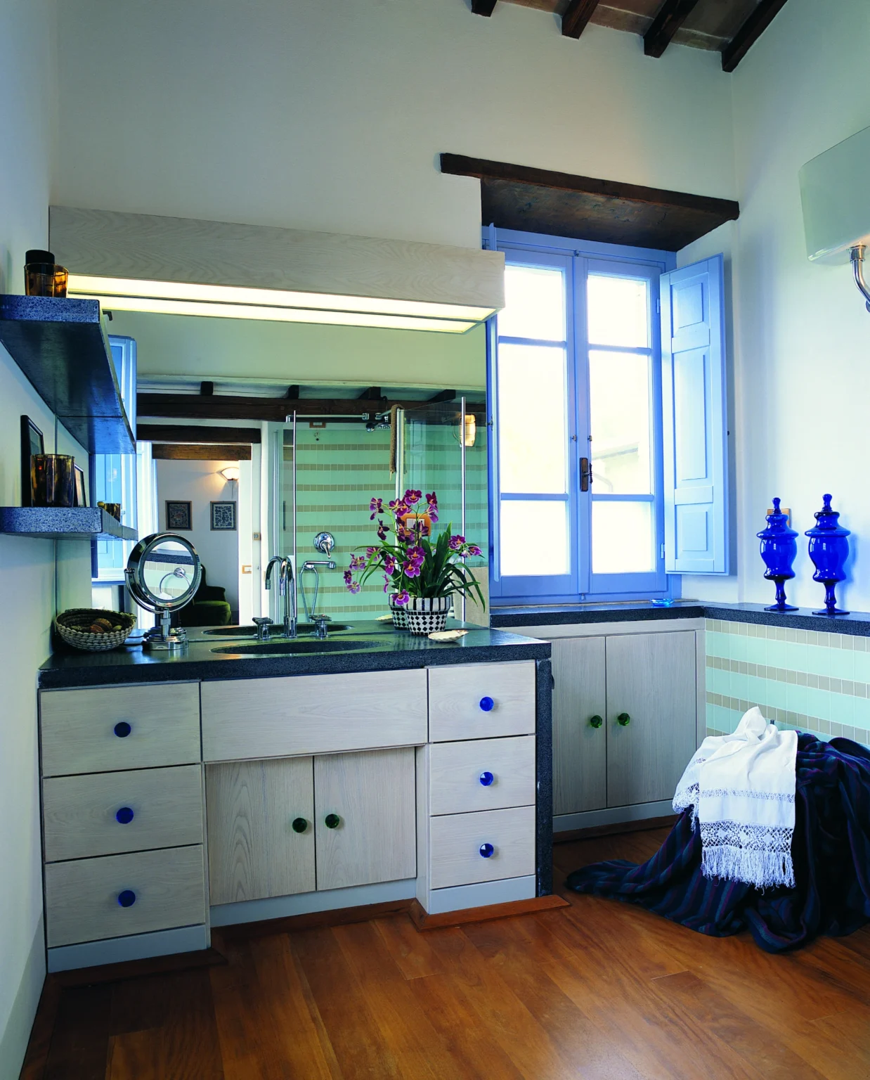Bright and modern kitchen interior featuring light green striped walls, a sleek dark countertop, and decorative blue glass vases. Natural light floods through a large window, enhancing the vibrant atmosphere. | Only on SopranoVillas