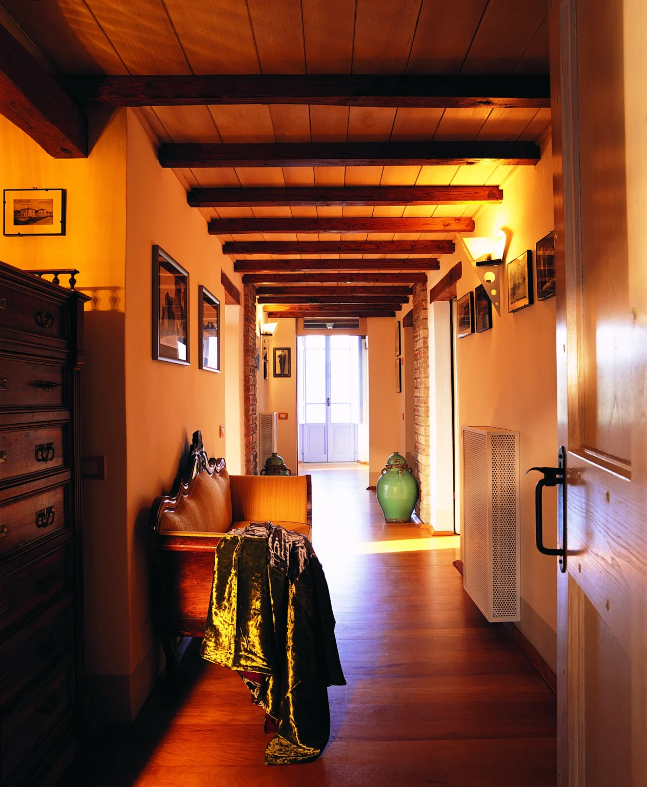 Cozy hallway with wooden beams, warm lighting, and decorative elements, featuring a vintage chest and a green vase, leading to a bright entrance. | Only on SopranoVillas