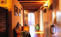 Cozy hallway with wooden beams, warm lighting, and decorative elements, featuring a vintage chest and a green vase, leading to a bright entrance.