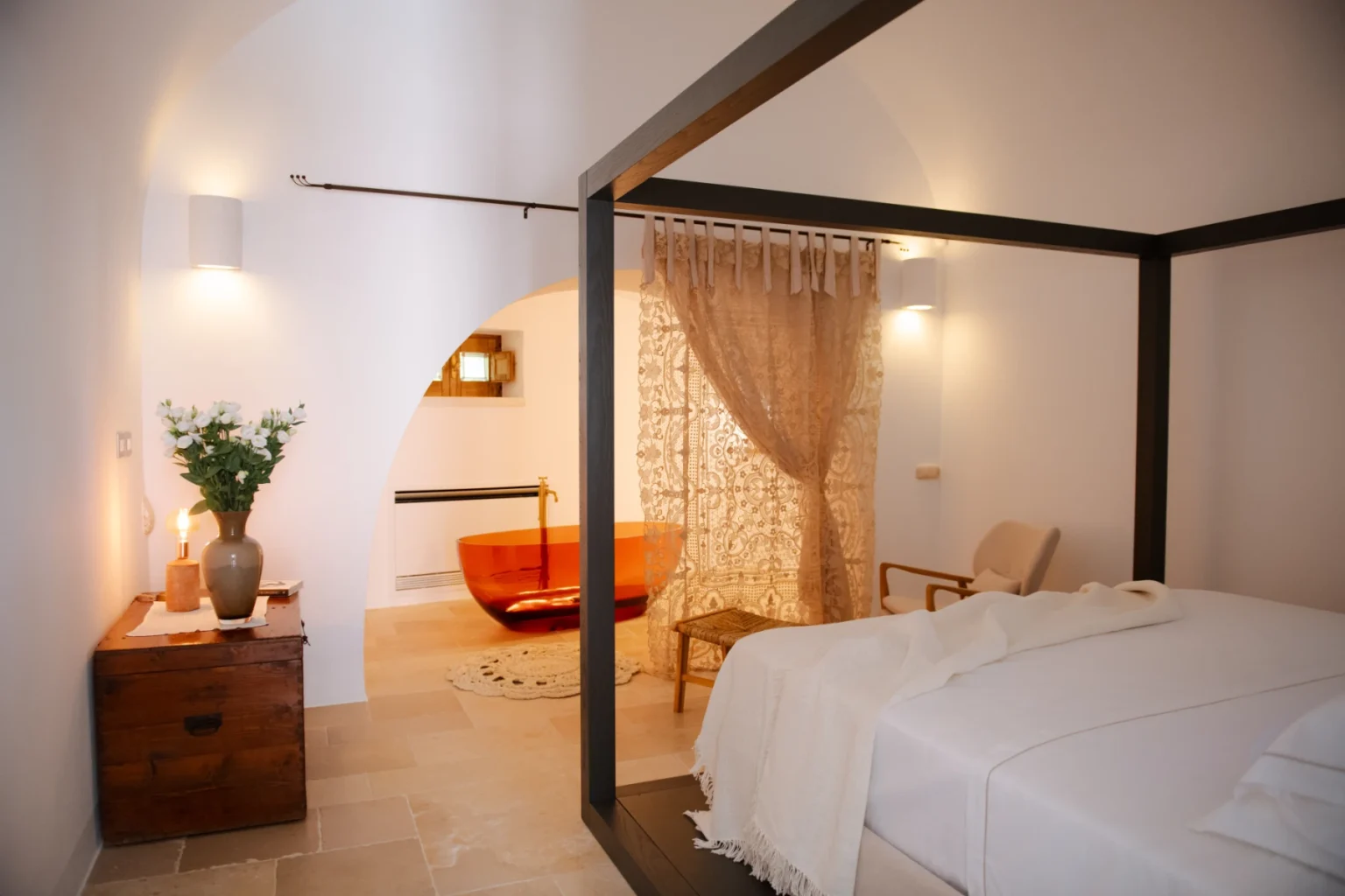 Modern bedroom interior featuring a four-poster bed, warm lighting, and minimalistic decor. The room includes a wooden nightstand, a cozy seating area, and elegant curtains, creating a serene atmosphere. | Only on SopranoVillas