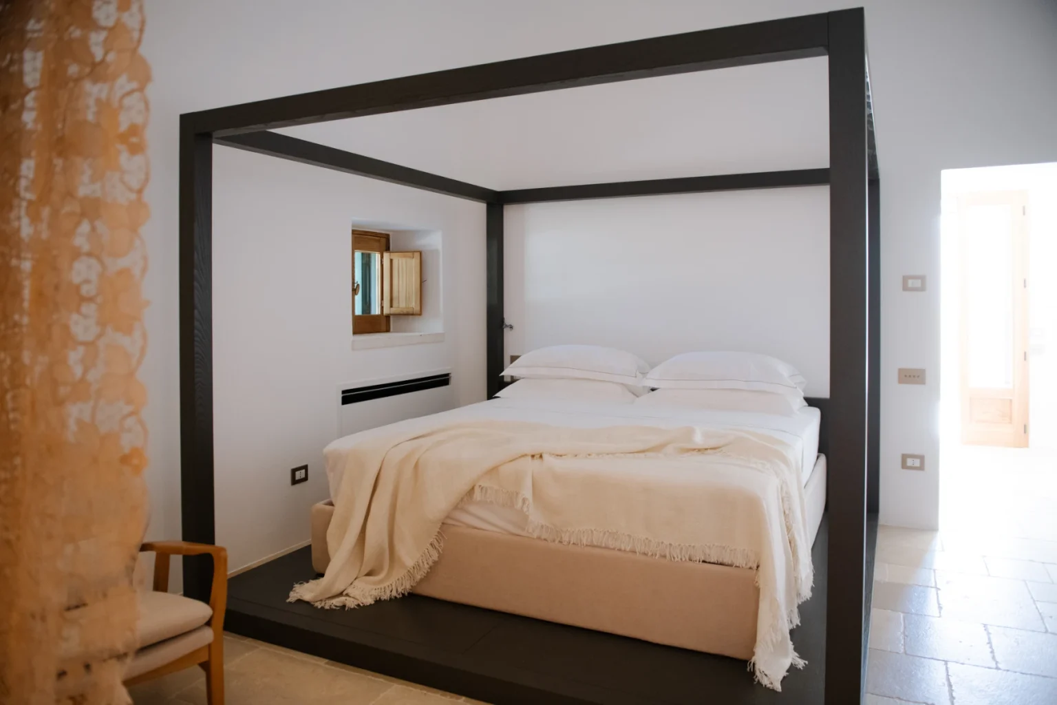 Modern bedroom featuring a stylish four-poster bed with white linens, minimalist decor, and natural light illuminating the space. | Only on SopranoVillas