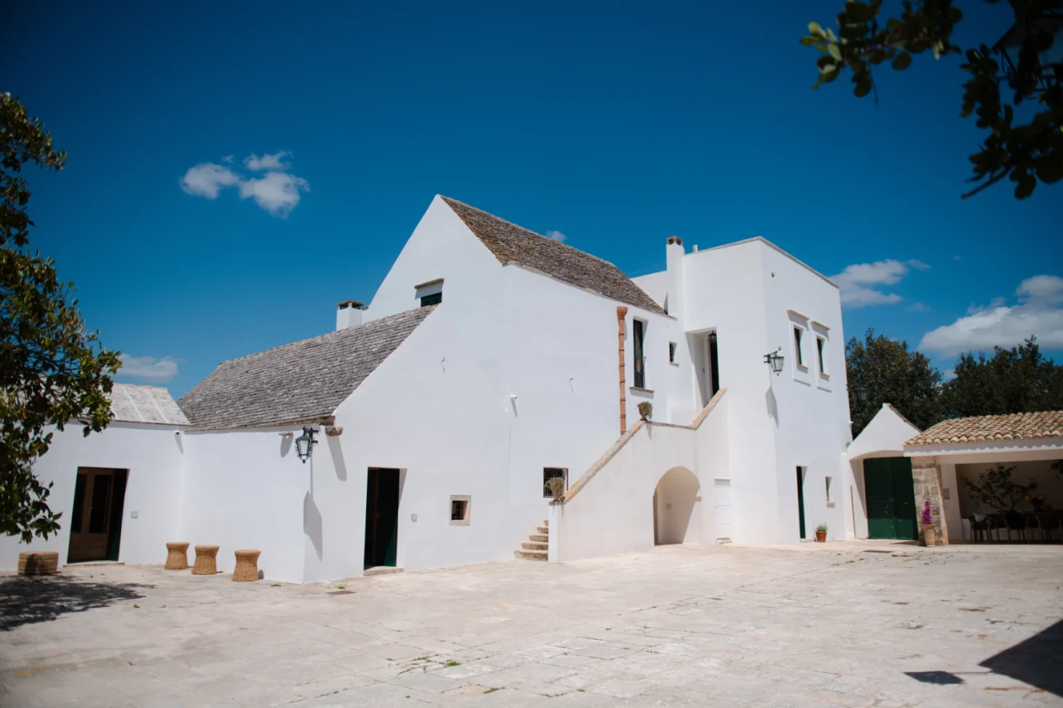 White traditional Italian villa with a stone roof, surrounded by trees and a clear blue sky. The building features a staircase leading to an upper level and a spacious courtyard. | Only on SopranoVillas