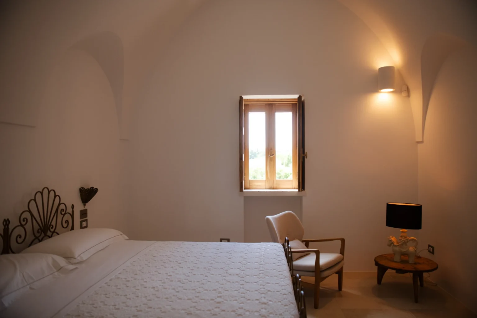 Cozy bedroom featuring a white bed with decorative headboard, a chair, a small side table, and a window with wooden shutters, illuminated by soft wall lighting. Ideal for relaxation and comfort. | Only on SopranoVillas