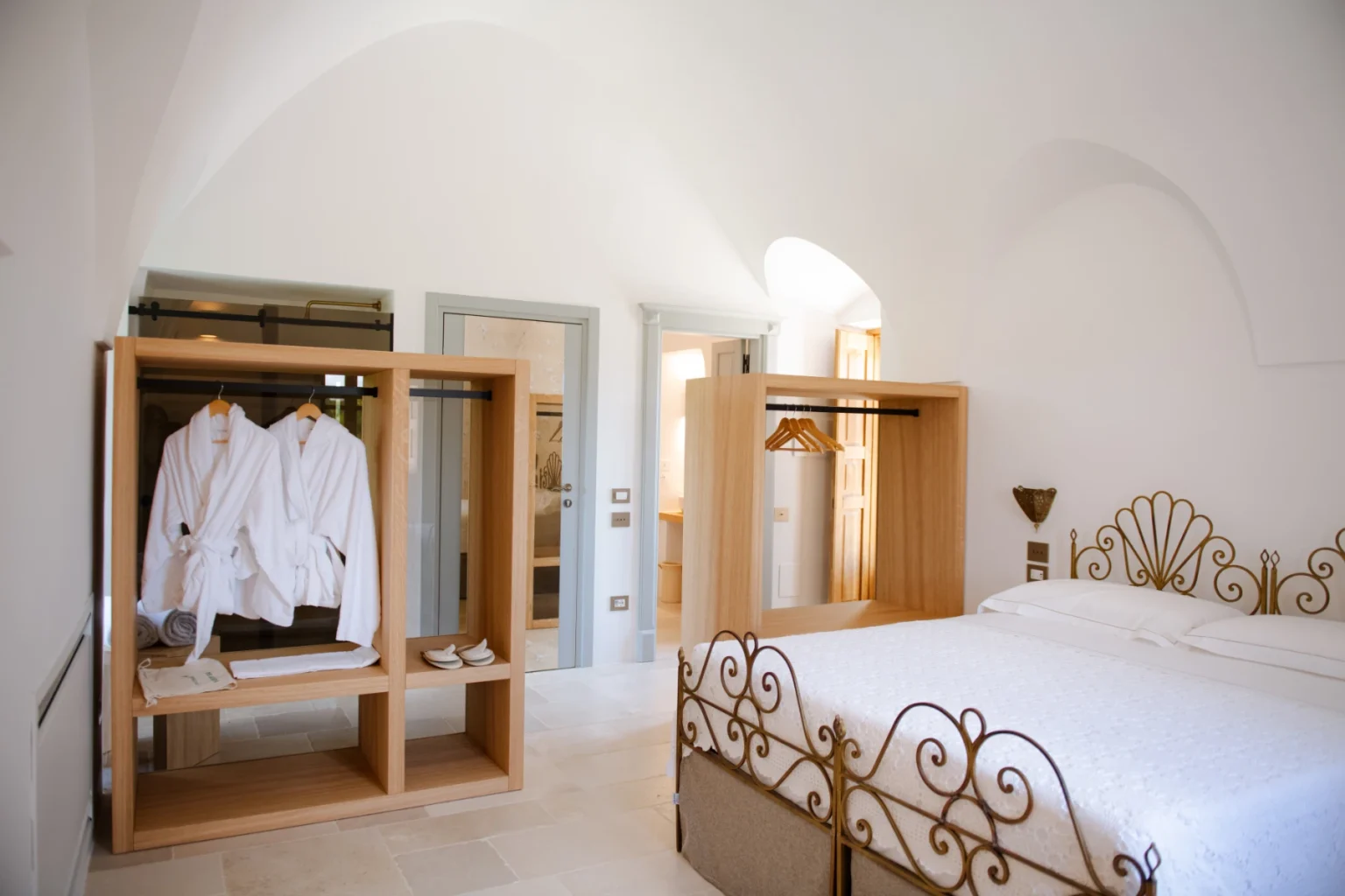 Bright and airy hotel room featuring a comfortable bed, elegant iron bed frame, and wooden wardrobe with neatly arranged white robes. The minimalist design includes natural light from arched windows, creating a serene atmosphere for relaxation. | Only on SopranoVillas