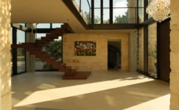 Modern interior of a spacious foyer featuring large glass walls, a wooden staircase, and contemporary artwork, illuminated by natural light.