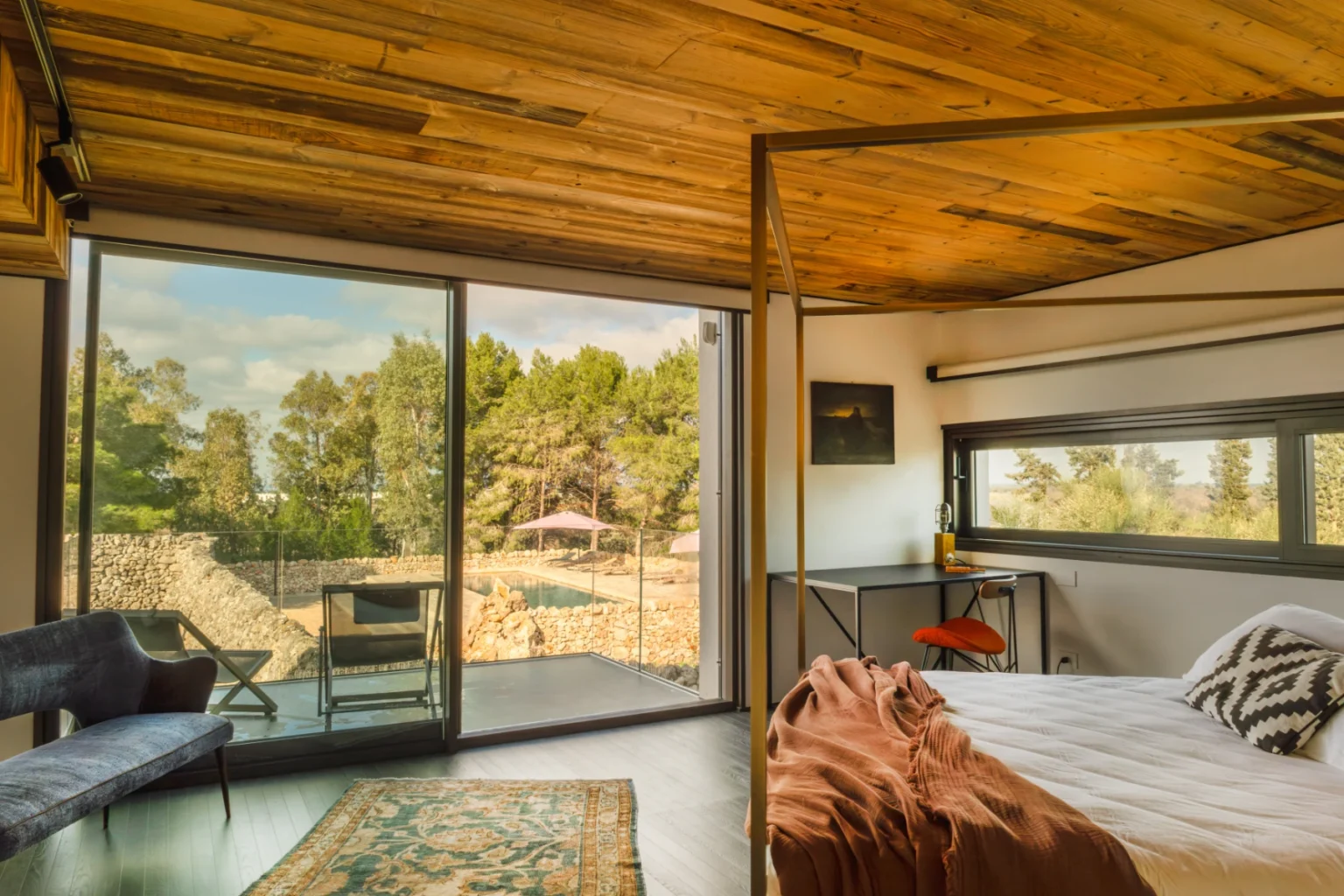 Modern bedroom with wooden ceiling, large glass doors leading to an outdoor view of trees and a pool, featuring a cozy bed, stylish decor, and a comfortable seating area. | Only on SopranoVillas