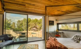 Modern bedroom with wooden ceiling, large glass doors leading to an outdoor view of trees and a pool, featuring a cozy bed, stylish decor, and a comfortable seating area.