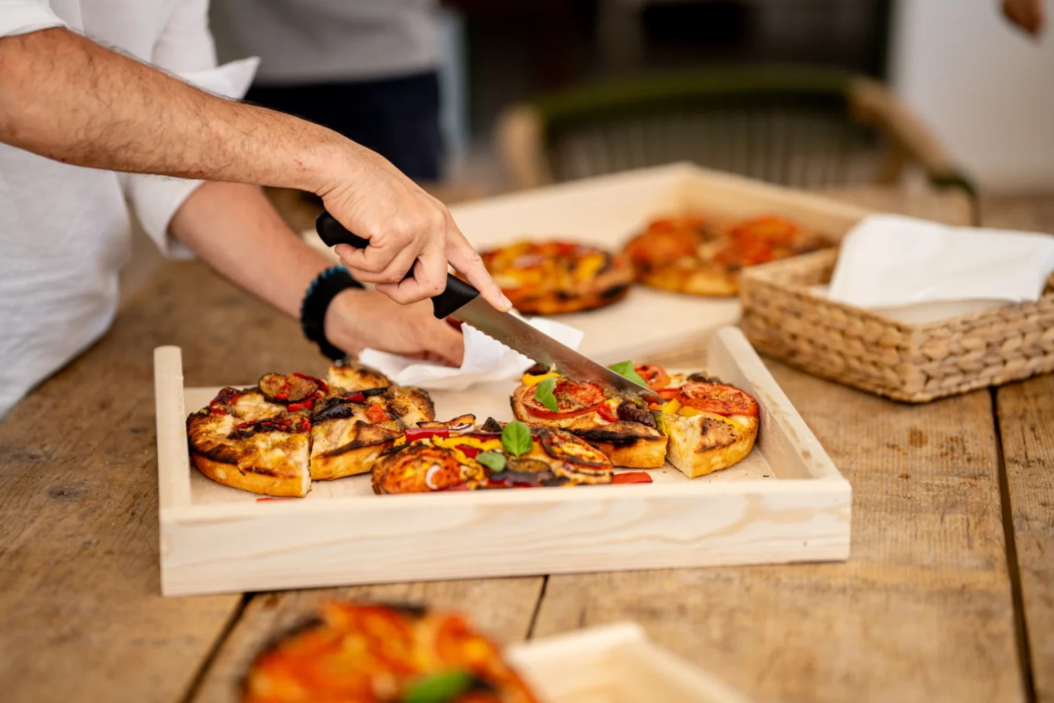 Person slicing a freshly baked pizza topped with vegetables on a wooden serving board, surrounded by additional pizzas and a rustic dining setting. | Only on SopranoVillas