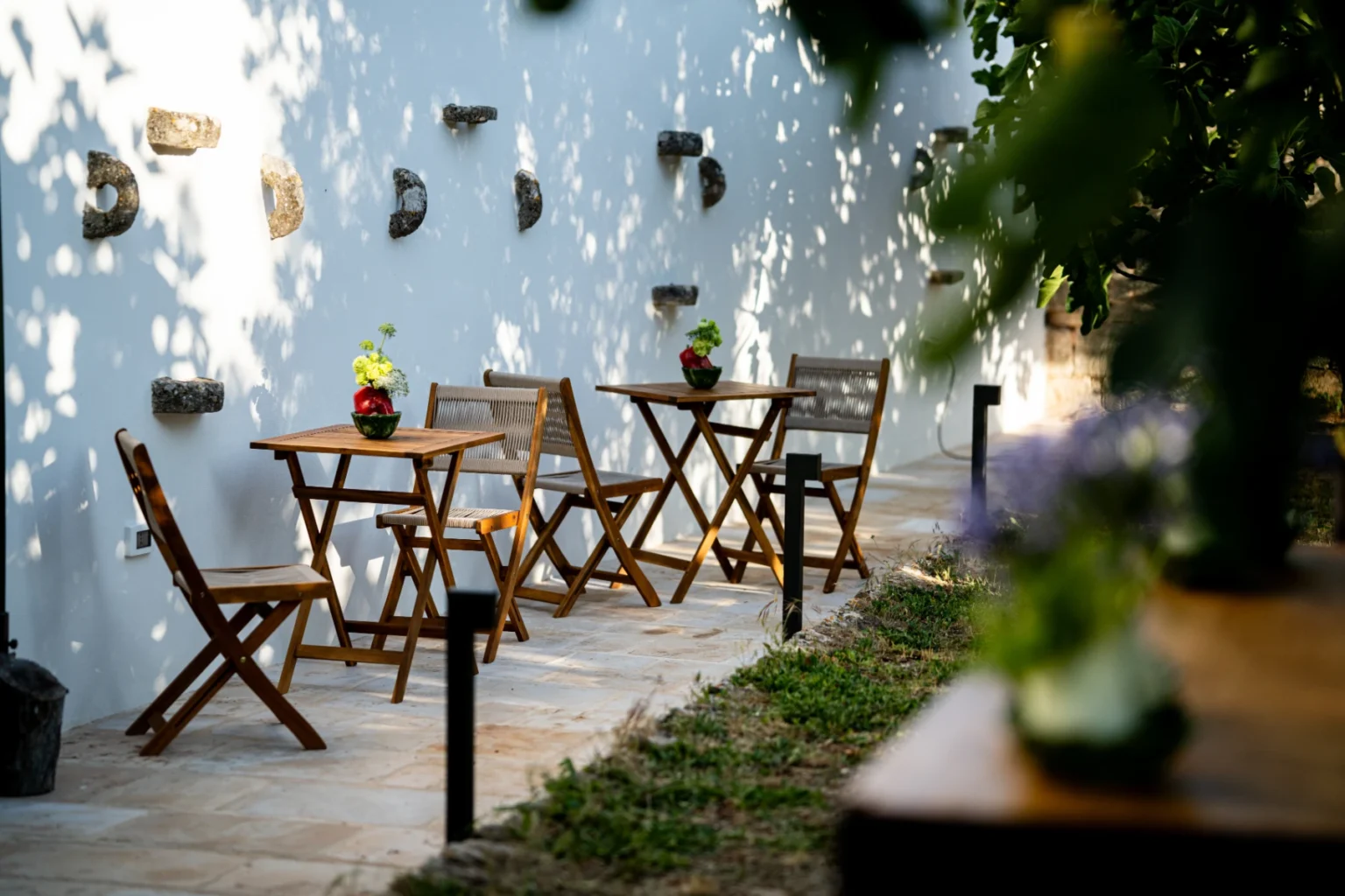 Outdoor café seating with wooden tables and chairs, adorned with potted plants, set against a white wall casting playful shadows. | Only on SopranoVillas