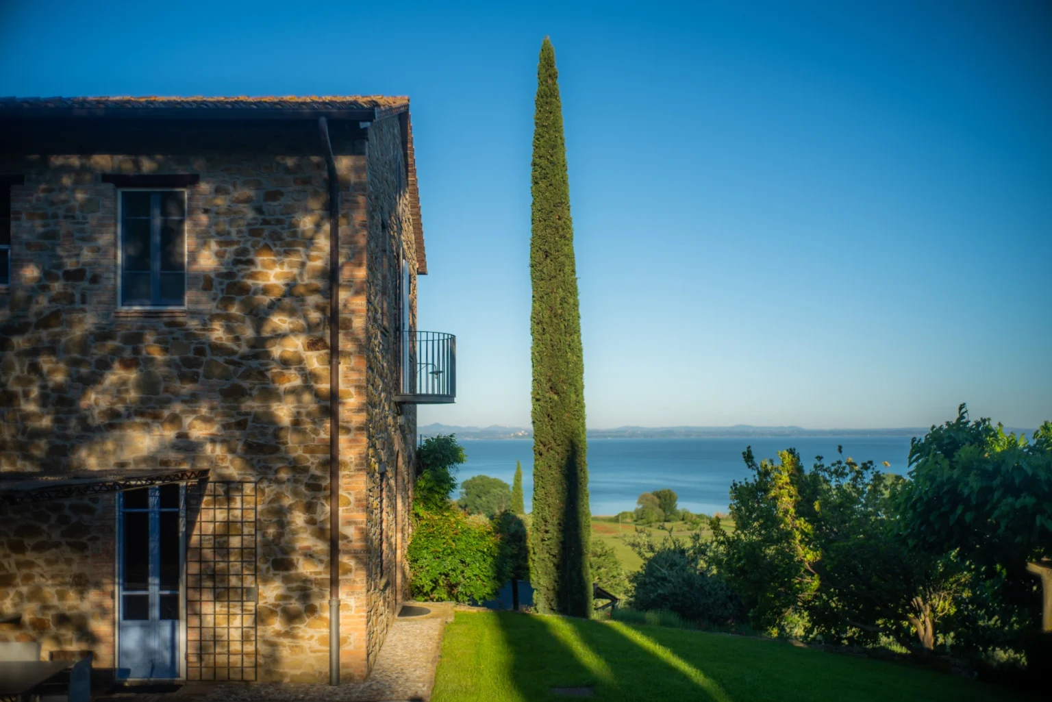 Scenic view of a rustic stone house beside a tall cypress tree, overlooking a tranquil sea and lush greenery under a clear blue sky. | Only on SopranoVillas