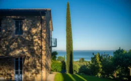 Scenic view of a rustic stone house beside a tall cypress tree, overlooking a tranquil sea and lush greenery under a clear blue sky.