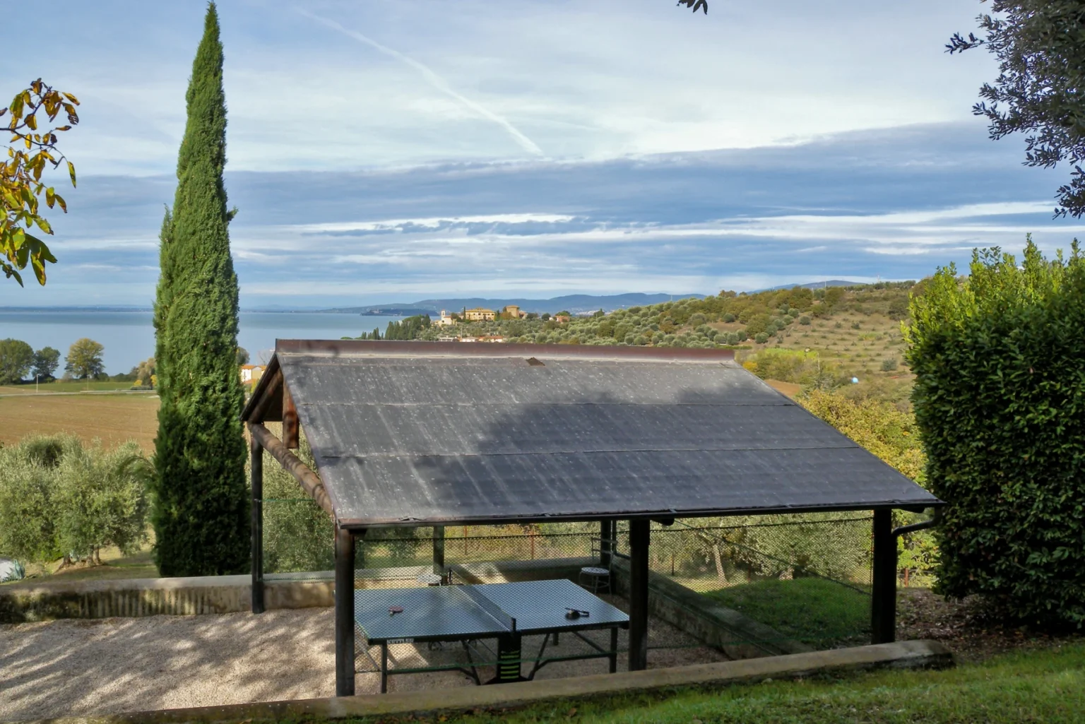 Scenic view of a wooden gazebo with a slate roof overlooking a tranquil landscape featuring a lake and rolling hills, surrounded by lush greenery and cypress trees. | Only on SopranoVillas