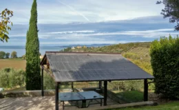 Scenic view of a wooden gazebo with a slate roof overlooking a tranquil landscape featuring a lake and rolling hills, surrounded by lush greenery and cypress trees.