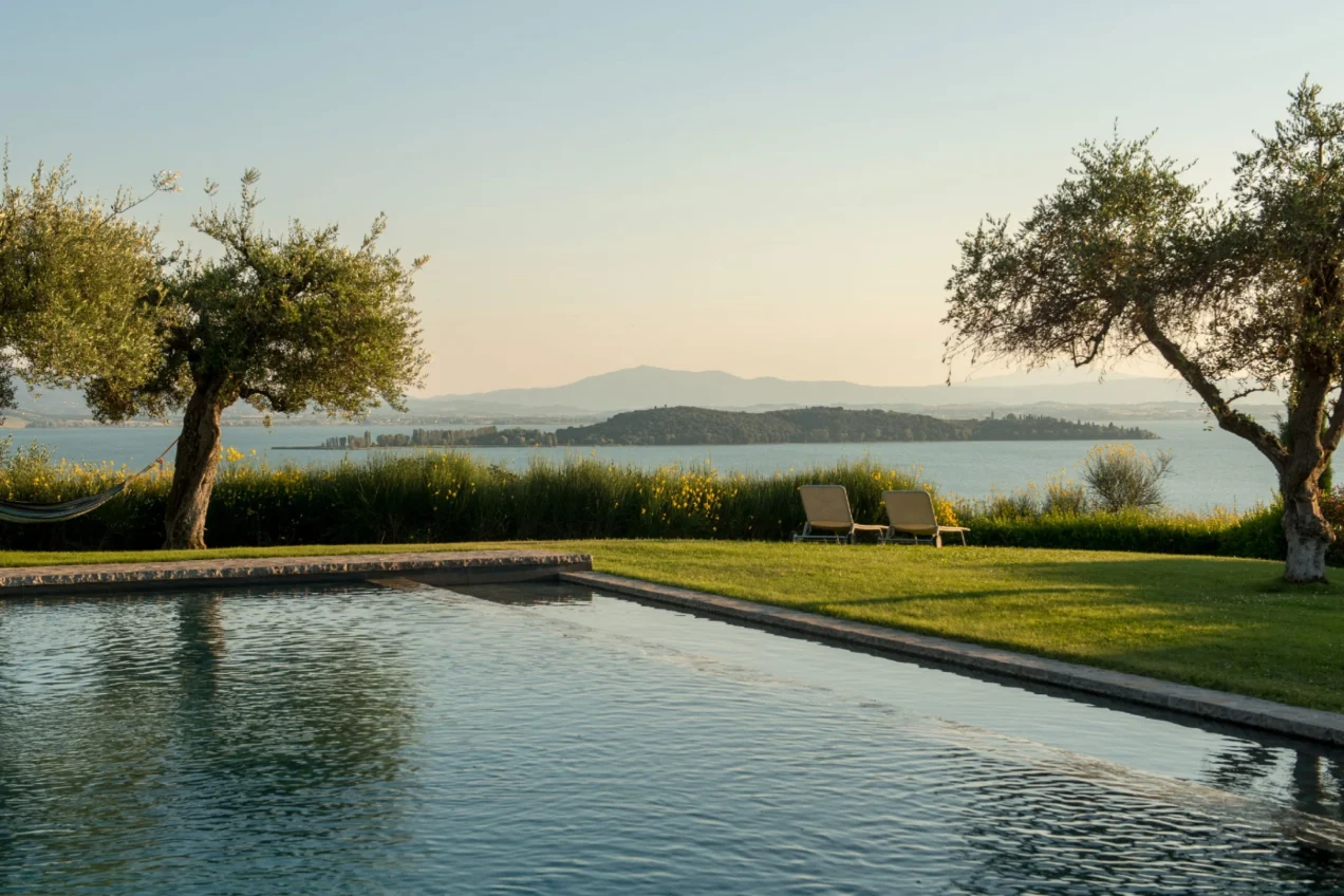 Scenic view of a tranquil swimming pool surrounded by lush greenery and olive trees, overlooking a serene lake and distant mountains at sunset. | Only on SopranoVillas