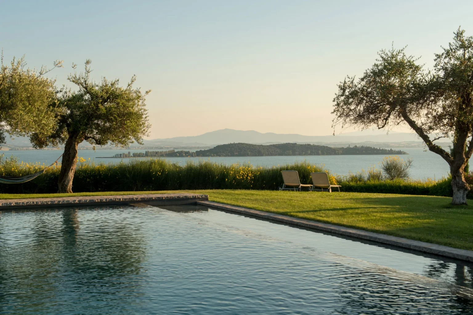 Scenic view of a tranquil swimming pool surrounded by lush greenery and olive trees, overlooking a serene lake and distant mountains at sunset. | Only on SopranoVillas