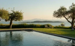 Scenic view of a tranquil swimming pool surrounded by lush greenery and olive trees, overlooking a serene lake and distant mountains at sunset.