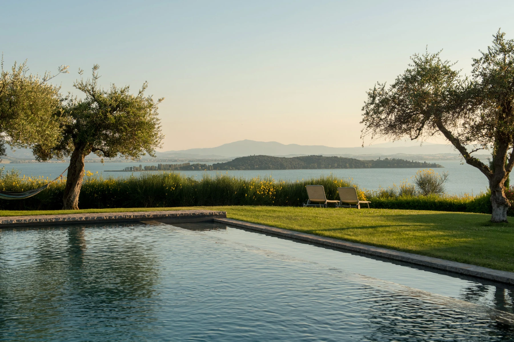 Scenic view of a tranquil swimming pool surrounded by lush greenery and olive trees, overlooking a serene lake and distant mountains at sunset. | Only on SopranoVillas
