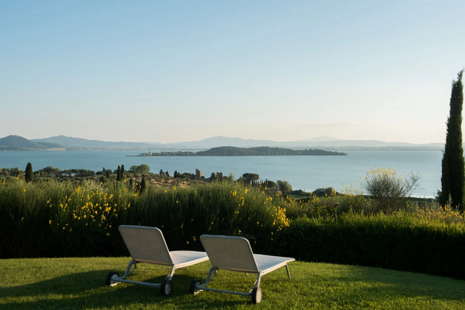 Scenic view of a tranquil lake surrounded by lush greenery, featuring two lounge chairs on a grassy area, perfect for relaxation and enjoying the serene landscape. | Only on SopranoVillas