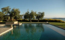 Luxury outdoor pool with clear blue water, surrounded by olive trees and lush greenery, offering a scenic view of the landscape and sky. Ideal for relaxation and leisure.