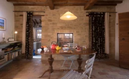 Cozy dining area featuring a wooden table set with colorful dishes, surrounded by rustic stone walls and warm wooden beams. Natural light illuminates the space, enhancing the inviting atmosphere.