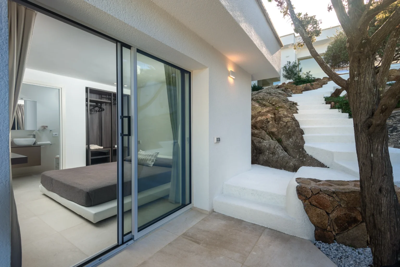 Modern bedroom with large glass doors opening to an outdoor space, featuring natural stone elements and a staircase leading up. The interior is minimalistic with neutral tones, creating a serene atmosphere. | Only on SopranoVillas