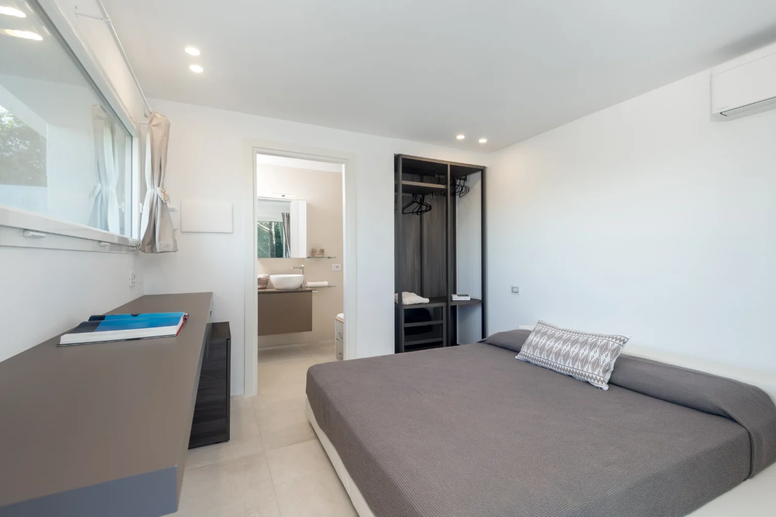 Modern bedroom with a gray bed, minimalist decor, and an ensuite bathroom. Features include a sleek dresser, open closet space, and ample natural light. | Only on SopranoVillas