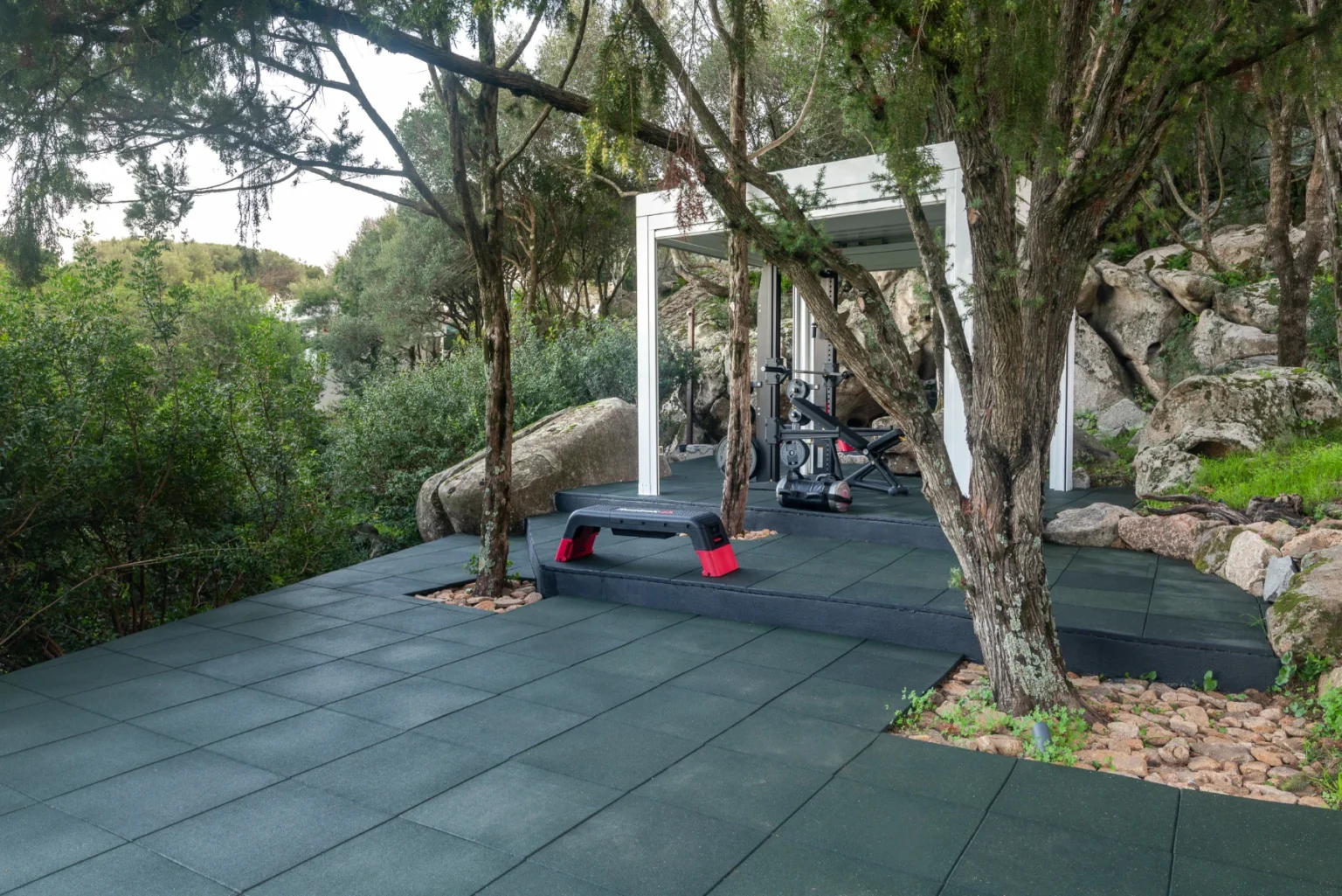 Outdoor home gym setup surrounded by greenery, featuring rubber flooring, exercise equipment, and a shaded area under a pergola. | Only on SopranoVillas