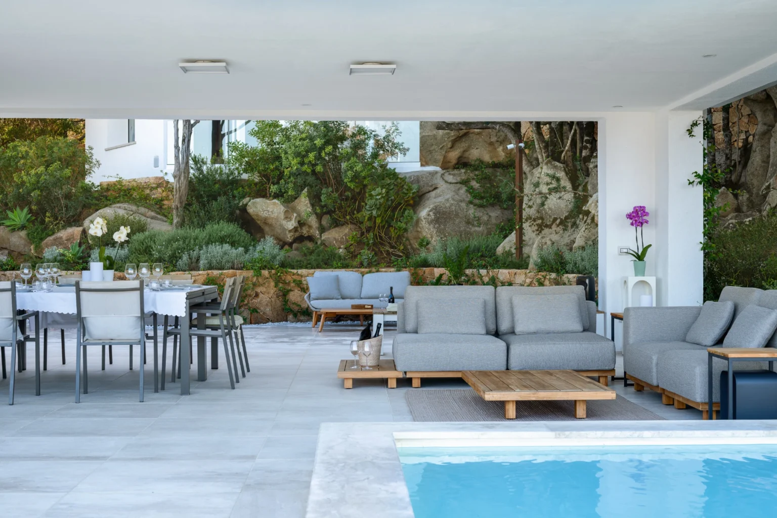 Modern outdoor living space featuring a sleek pool, comfortable gray sofas, and a dining area with a stylish table, surrounded by lush greenery and natural stone elements. Ideal for relaxation and entertaining. | Only on SopranoVillas