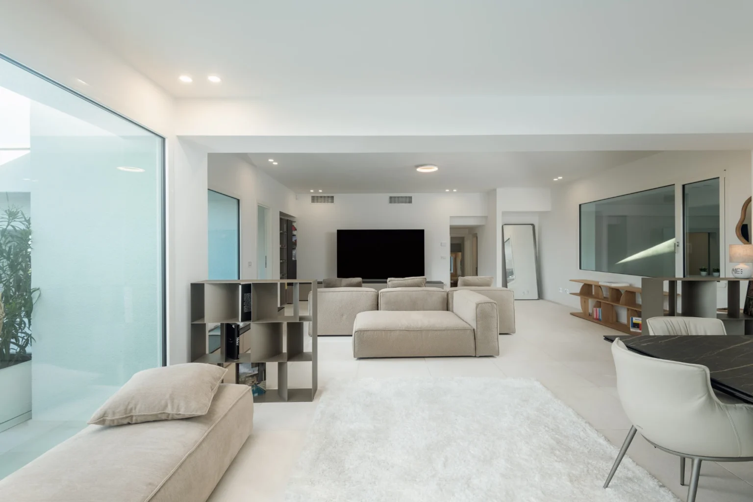 Modern living room featuring neutral-toned sofas, a large flat-screen TV, and glass walls that enhance natural light. The minimalist design includes a stylish shelving unit and a sleek dining area, creating an open and inviting space. | Only on SopranoVillas