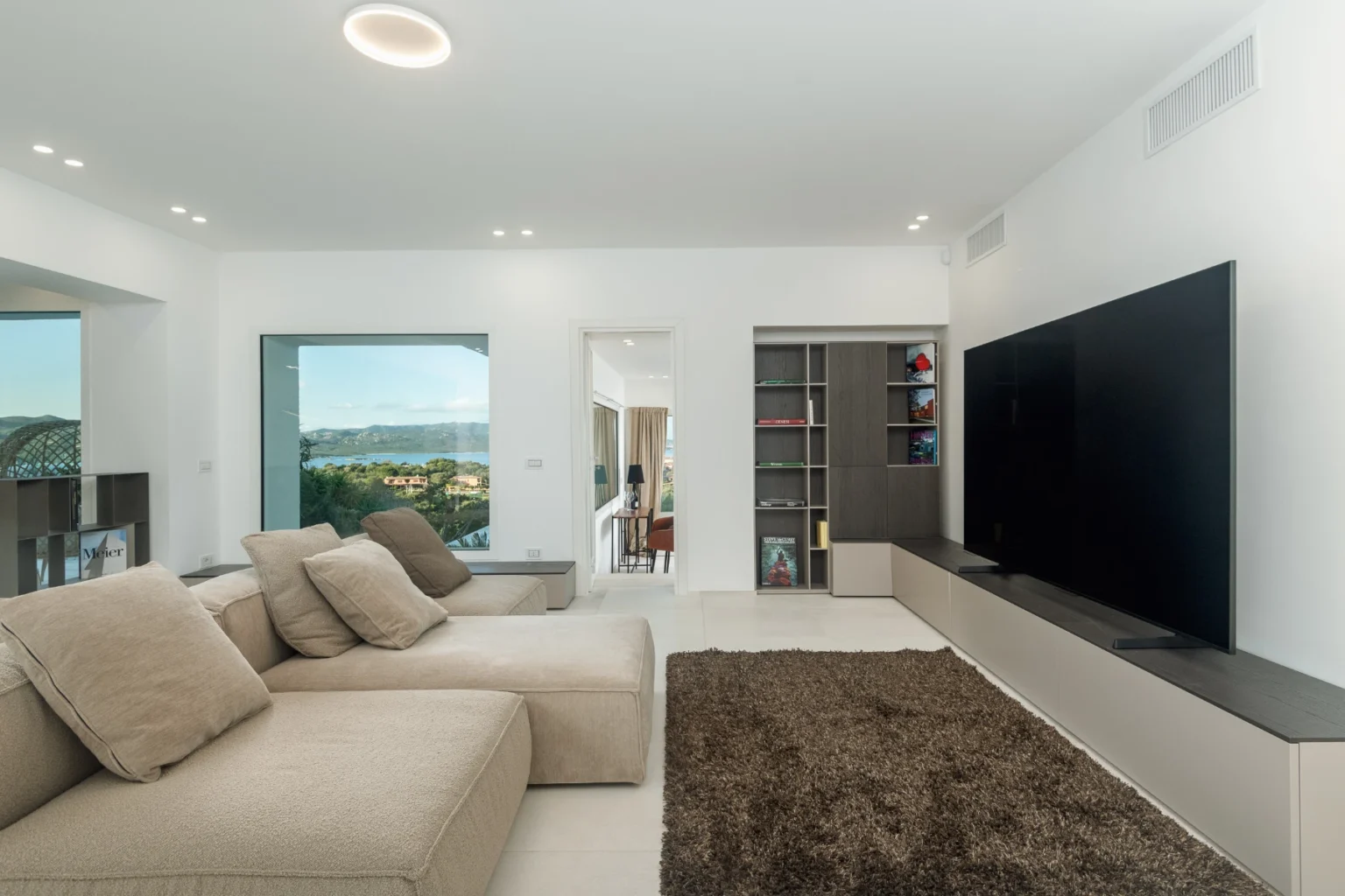 Modern living room featuring a large flat-screen TV, a sectional beige sofa, and a plush area rug. Bright natural light streams in through large windows, highlighting the minimalist decor and contemporary bookshelf. Ideal for showcasing stylish interior design and comfortable living spaces. | Only on SopranoVillas