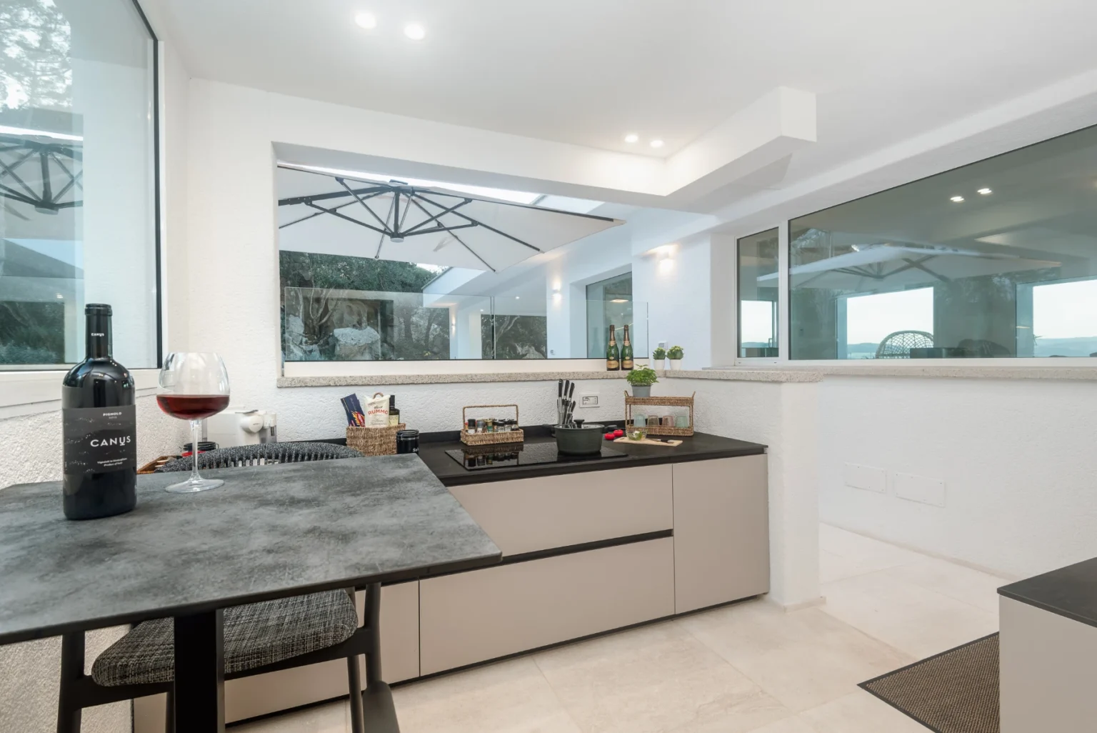 Modern kitchen with sleek beige cabinetry, a black countertop, and large windows offering a view of the outdoors. A wine bottle and glass are prominently displayed on the counter, enhancing the inviting atmosphere. | Only on SopranoVillas