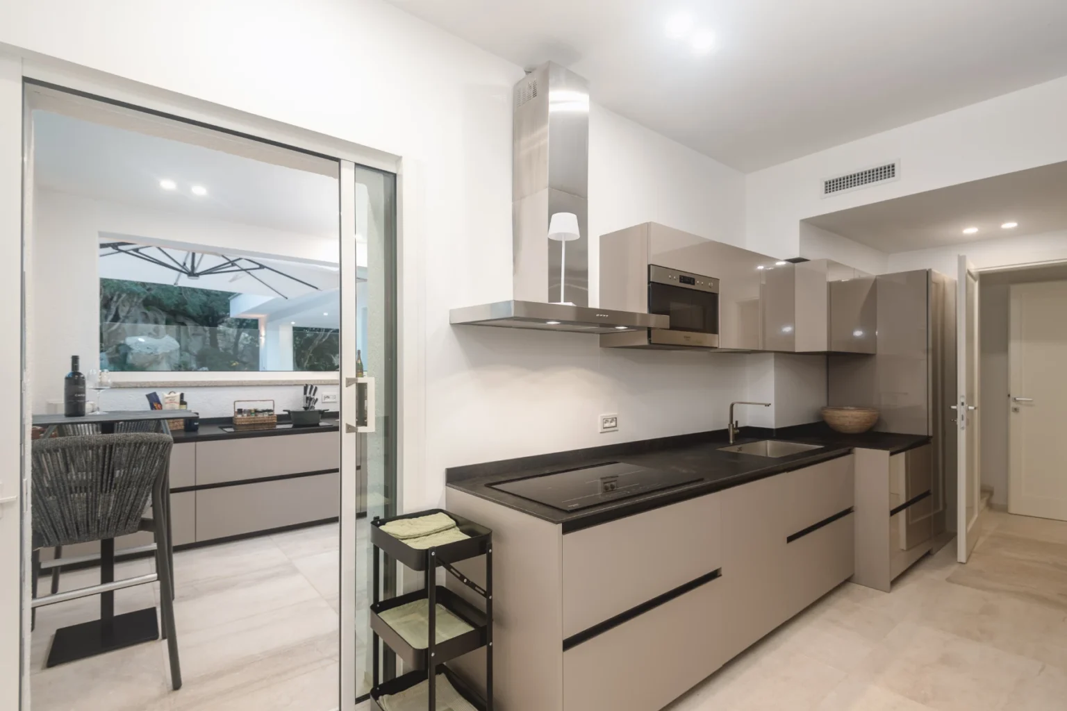 Modern kitchen interior featuring sleek gray cabinets, a black countertop, and stainless steel appliances, with an open layout leading to a dining area. | Only on SopranoVillas