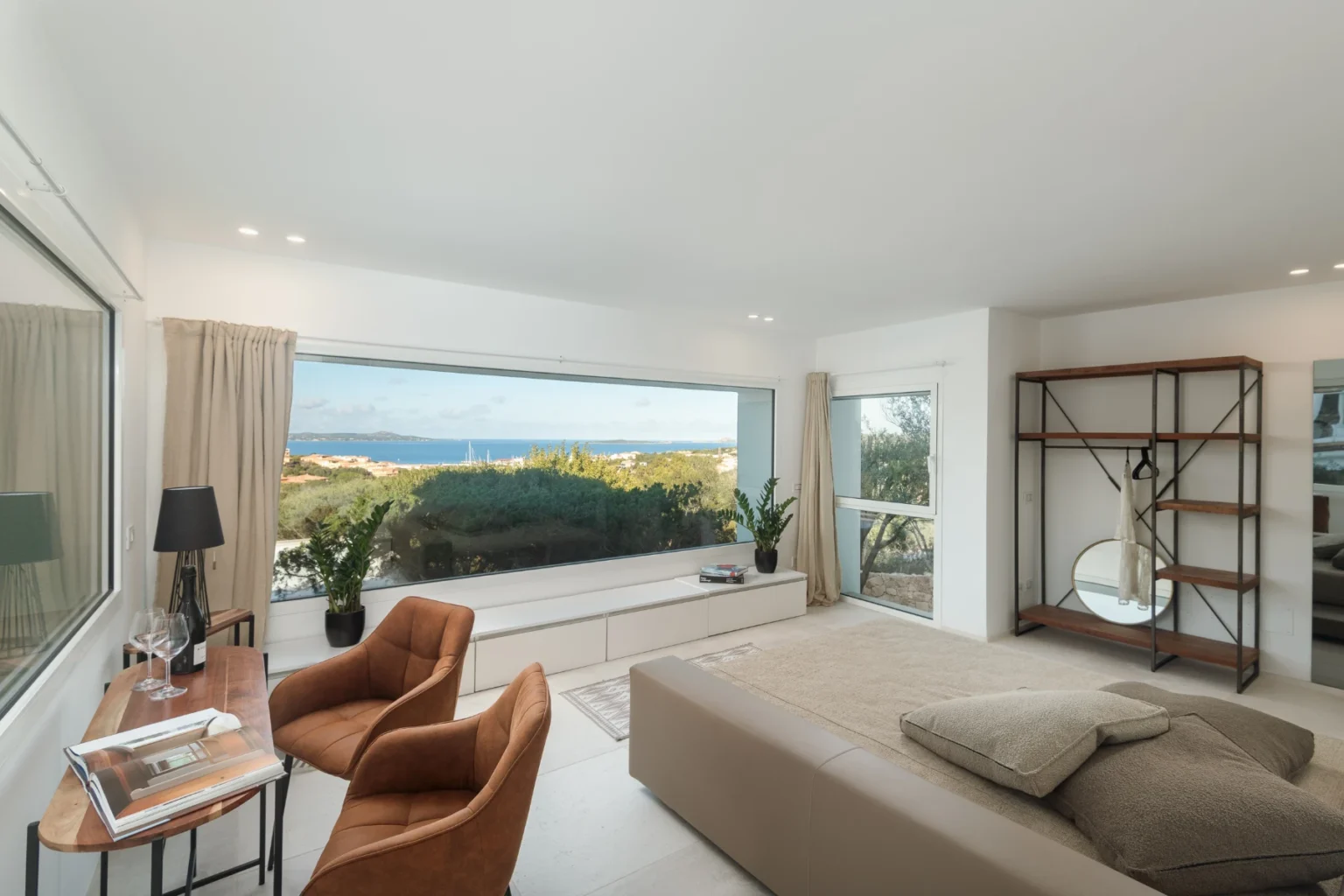 Modern bedroom with large windows offering a scenic ocean view, featuring a cozy bed, stylish armchairs, and a wooden shelf, designed for comfort and relaxation. | Only on SopranoVillas
