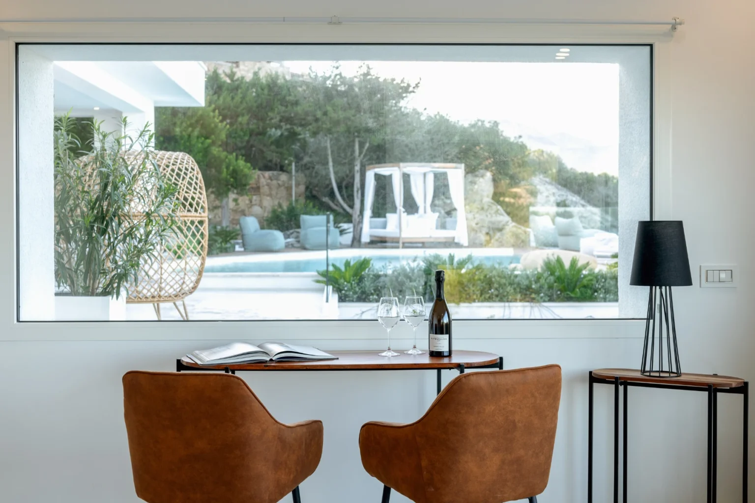 Cozy indoor seating area featuring two brown chairs and a wooden table, overlooking a serene outdoor pool and garden through a large window. A bottle of wine and glasses are placed on the table, accompanied by a stylish lamp. | Only on SopranoVillas