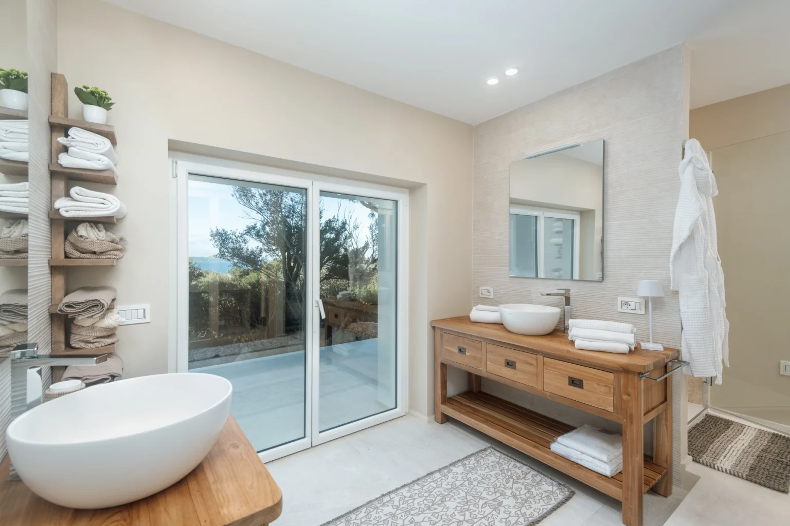 Modern bathroom interior featuring a stylish wooden vanity, a round vessel sink, and a large mirror. Shelving holds neatly stacked towels, while sliding glass doors provide natural light and a view of the outdoors. | Only on SopranoVillas