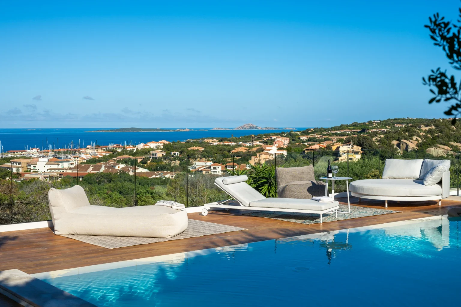 Luxury villa with a swimming pool overlooking a coastal town and the ocean, featuring modern outdoor furniture and a clear blue sky. | Only on SopranoVillas