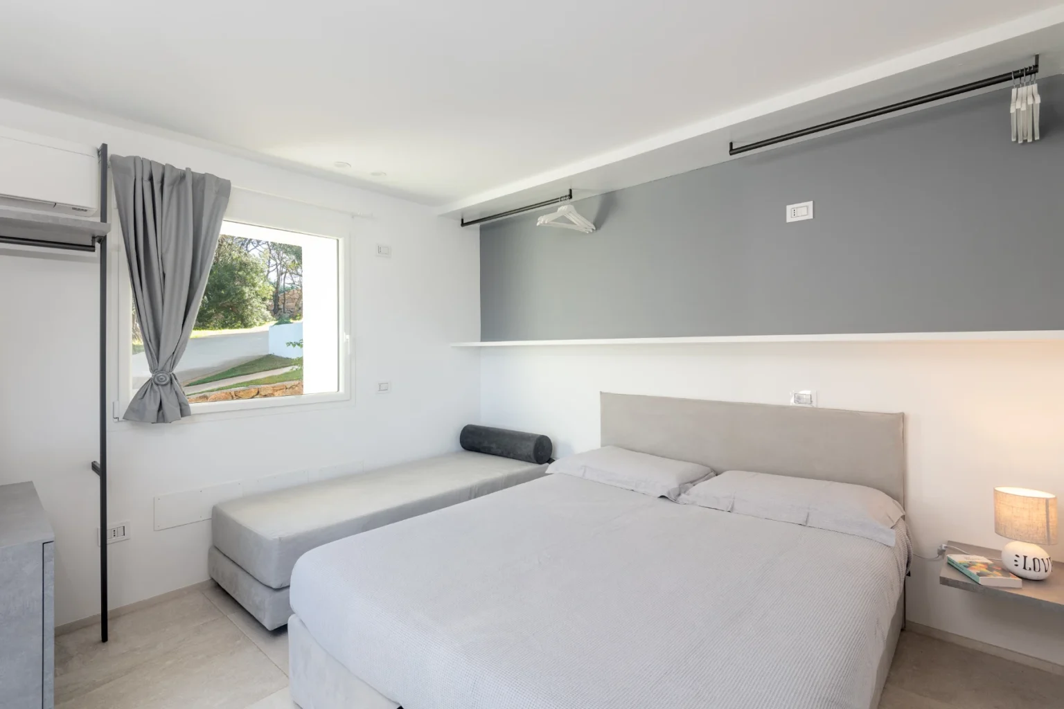 Modern bedroom featuring a queen-sized bed, a single bed, and a minimalist design with natural light from a window. The room includes gray walls, a nightstand with a lamp, and simple furnishings for a cozy atmosphere. | Only on SopranoVillas
