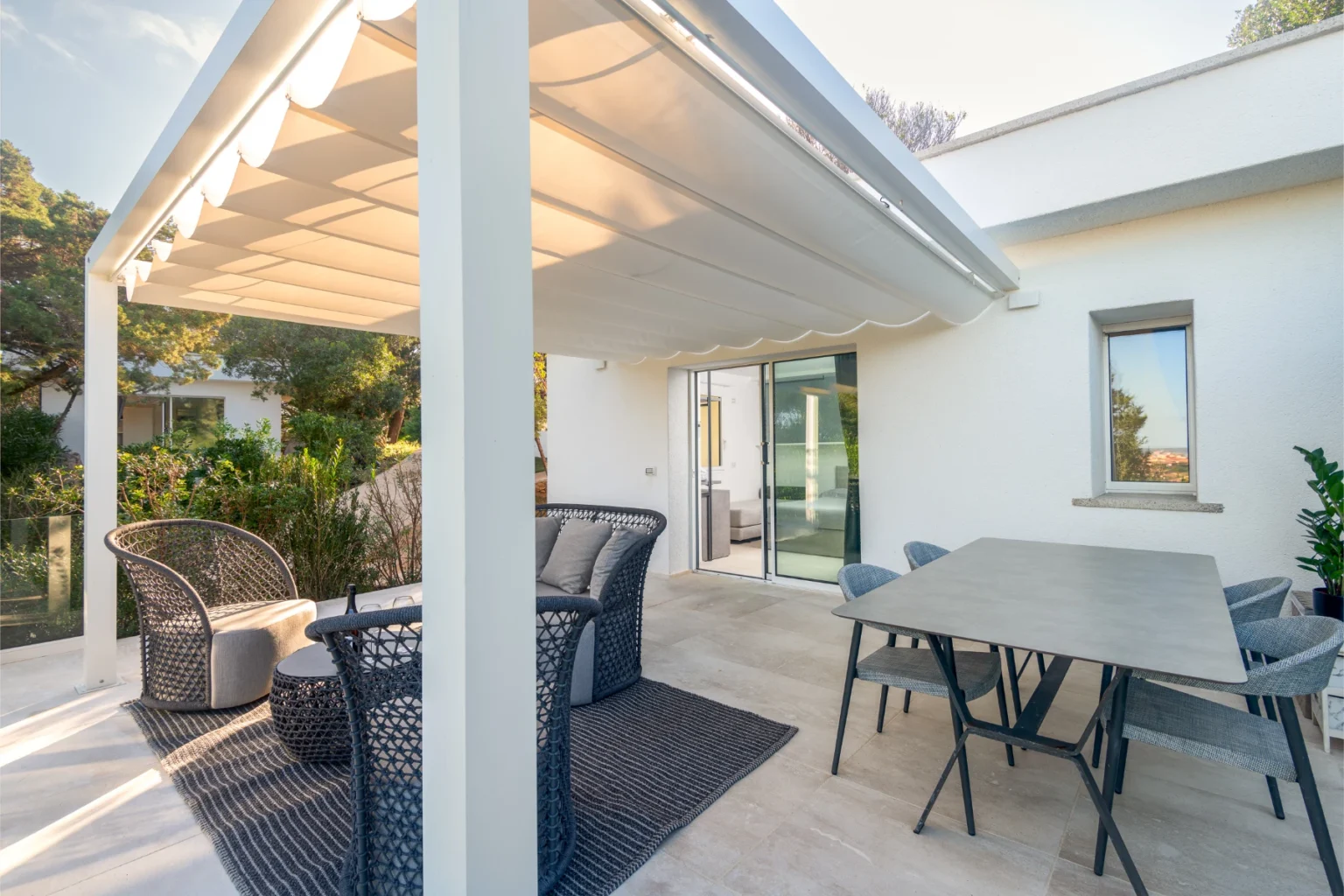 Modern outdoor patio featuring a stylish seating area with gray wicker furniture, a dining table, and a retractable awning, surrounded by greenery. | Only on SopranoVillas