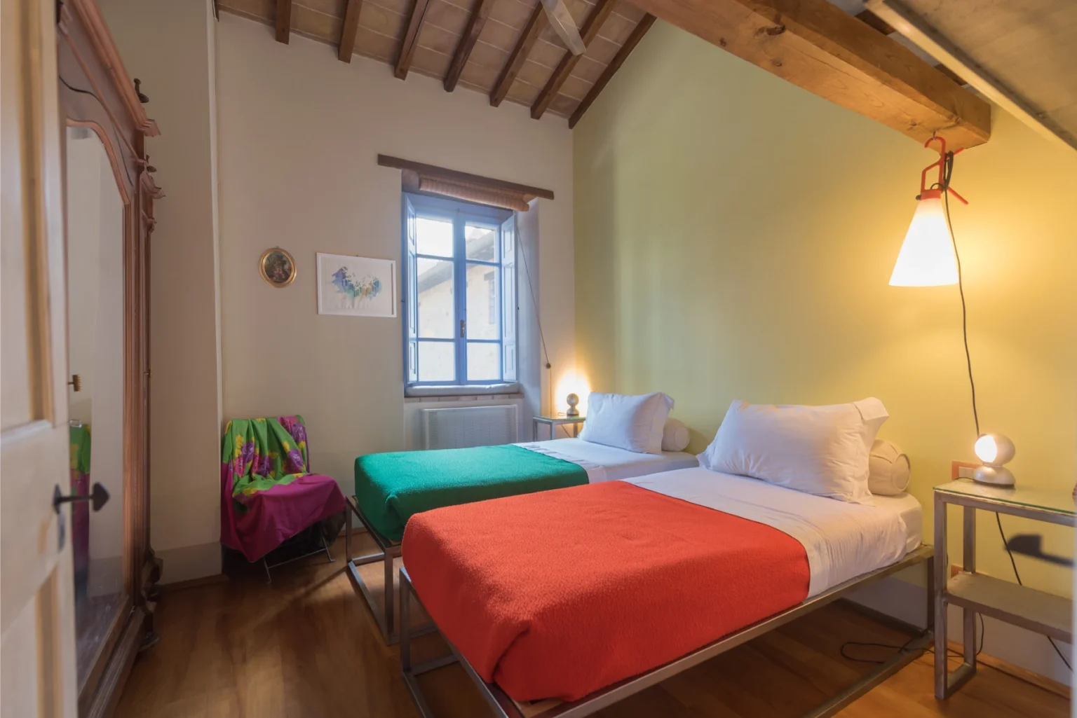 Cozy bedroom with two beds, one green and one orange, featuring wooden beams and a window that lets in natural light. The room includes a small bedside table and a colorful chair, creating a welcoming atmosphere. Perfect for a relaxing stay. | Only on SopranoVillas