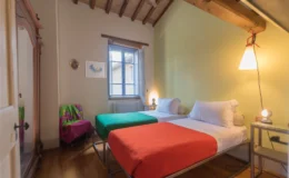 Cozy bedroom with two beds, one green and one orange, featuring wooden beams and a window that lets in natural light. The room includes a small bedside table and a colorful chair, creating a welcoming atmosphere. Perfect for a relaxing stay.