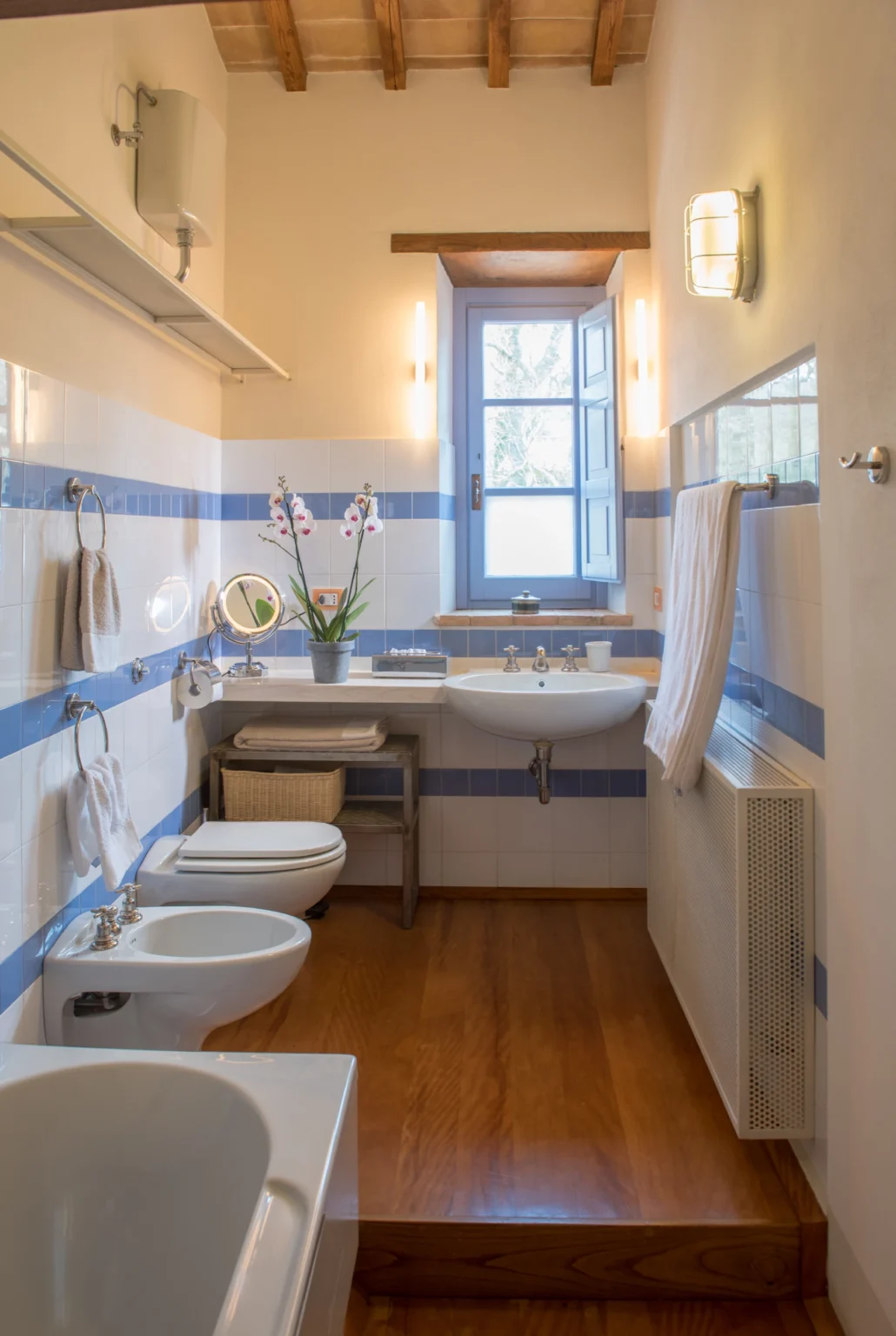 Bright and modern bathroom featuring blue and white striped walls, a sink with a mirror, a toilet, and a towel rack. Natural light enters through a window adorned with plants, creating a fresh and inviting atmosphere. | Only on SopranoVillas