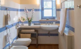 Bright and modern bathroom featuring blue and white striped walls, a sink with a mirror, a toilet, and a towel rack. Natural light enters through a window adorned with plants, creating a fresh and inviting atmosphere.