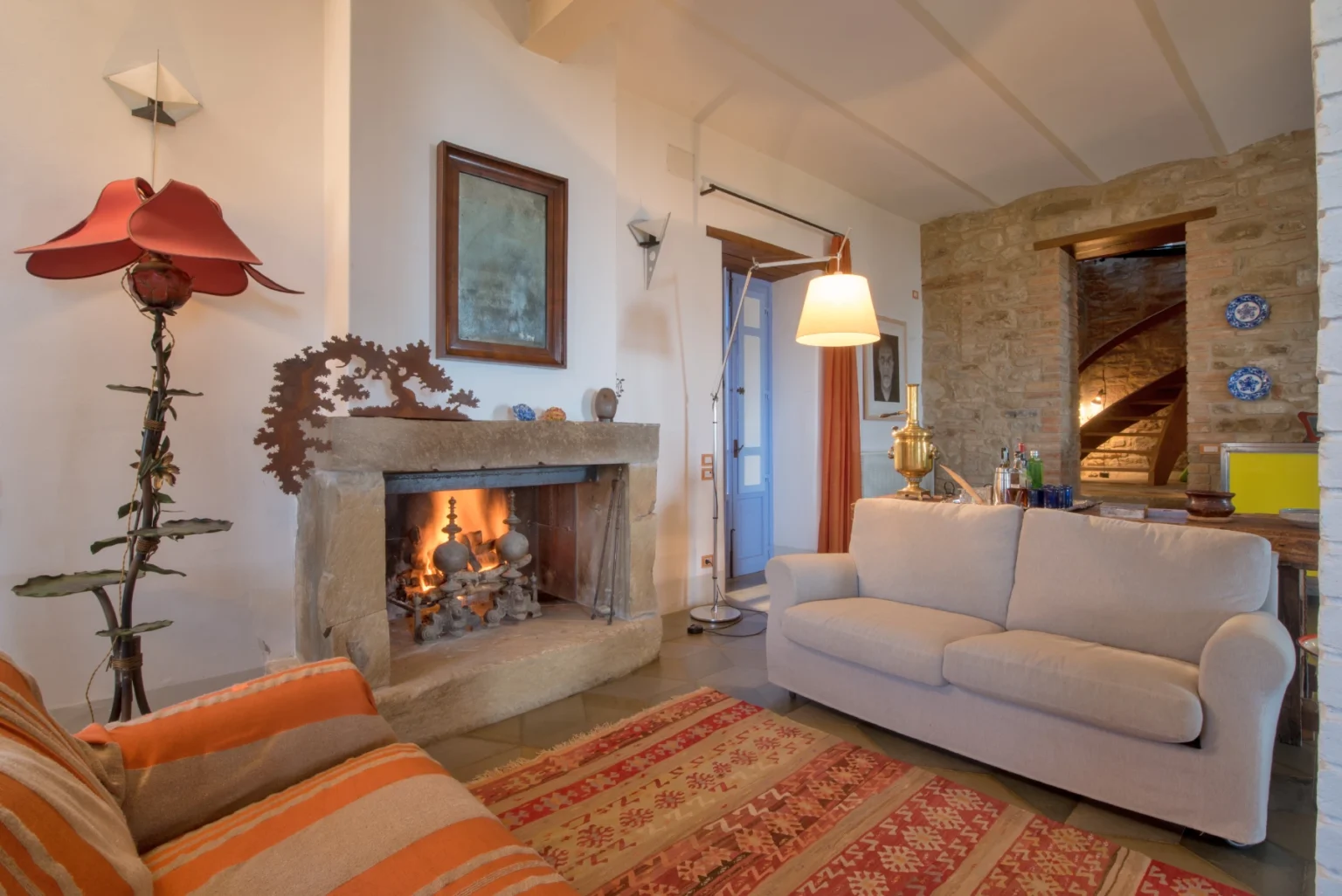 Cozy living room featuring a stone fireplace, a comfortable beige sofa, and warm lighting. The decor includes a colorful area rug and a unique floor lamp, creating an inviting atmosphere. | Only on SopranoVillas