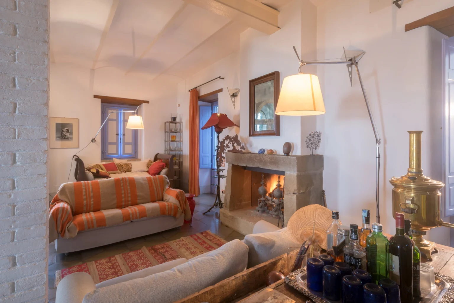 Cozy living room featuring a comfortable sofa with colorful throw pillows, a stone fireplace, and warm lighting. The decor includes artwork on the walls and a bar cart with various bottles, creating an inviting atmosphere. | Only on SopranoVillas